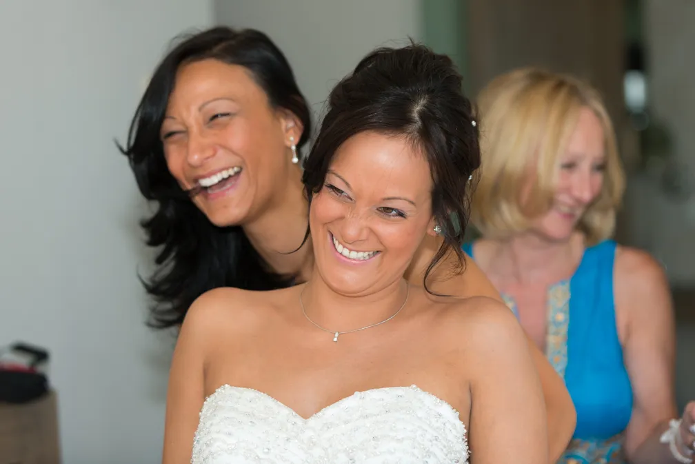 Bride in a white strapless wedding dress smiling with two women laughing behind her.