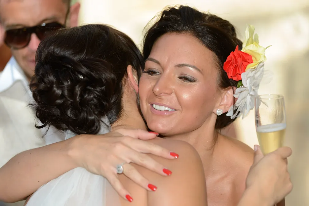 Two women embracing, one holding a champagne glass and wearing red and yellow flowers in her hair.
