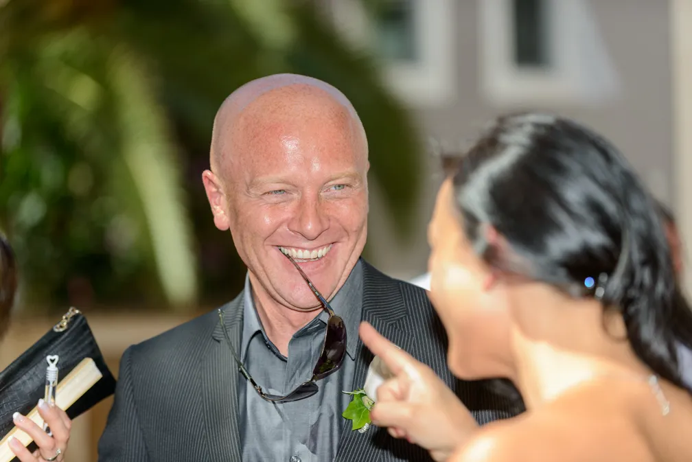 Smiling bald man in a suit holding sunglasses in his mouth engaging with a woman pointing at him.