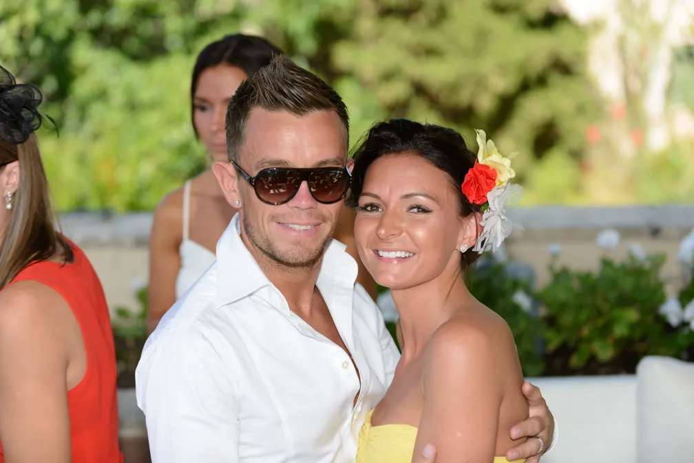 Smiling couple embracing at an outdoor event, the man wearing sunglasses and a white shirt, the woman wearing a yellow dress and floral hair accessories.