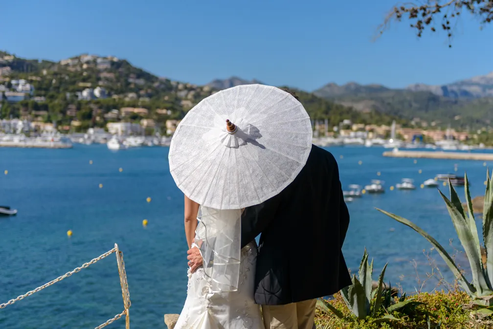 A couple embraces while holding a white parasol, overlooking a coastal town with boats on blue water and mountains in the background.