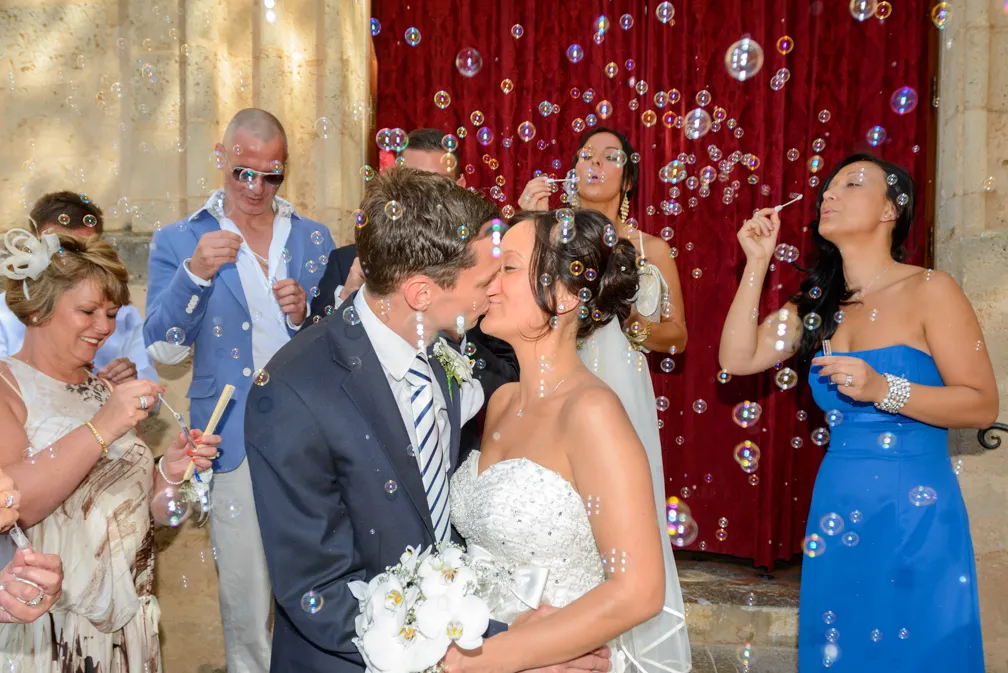 Bride and groom kissing surrounded by guests blowing bubbles at a wedding celebration.