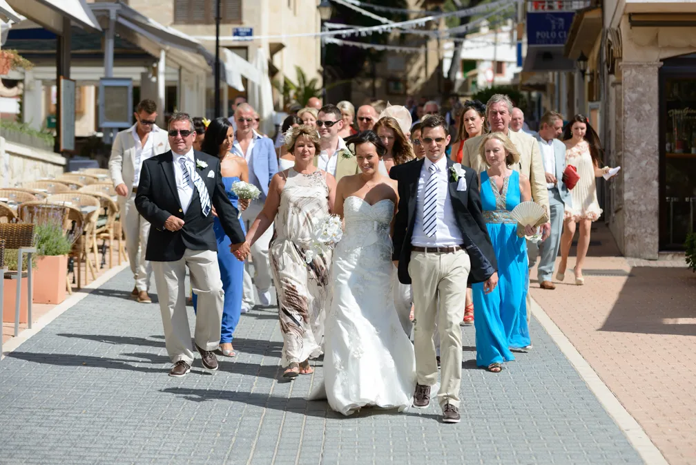 Bride in white wedding dress walking with groom and guests on a sunny street.