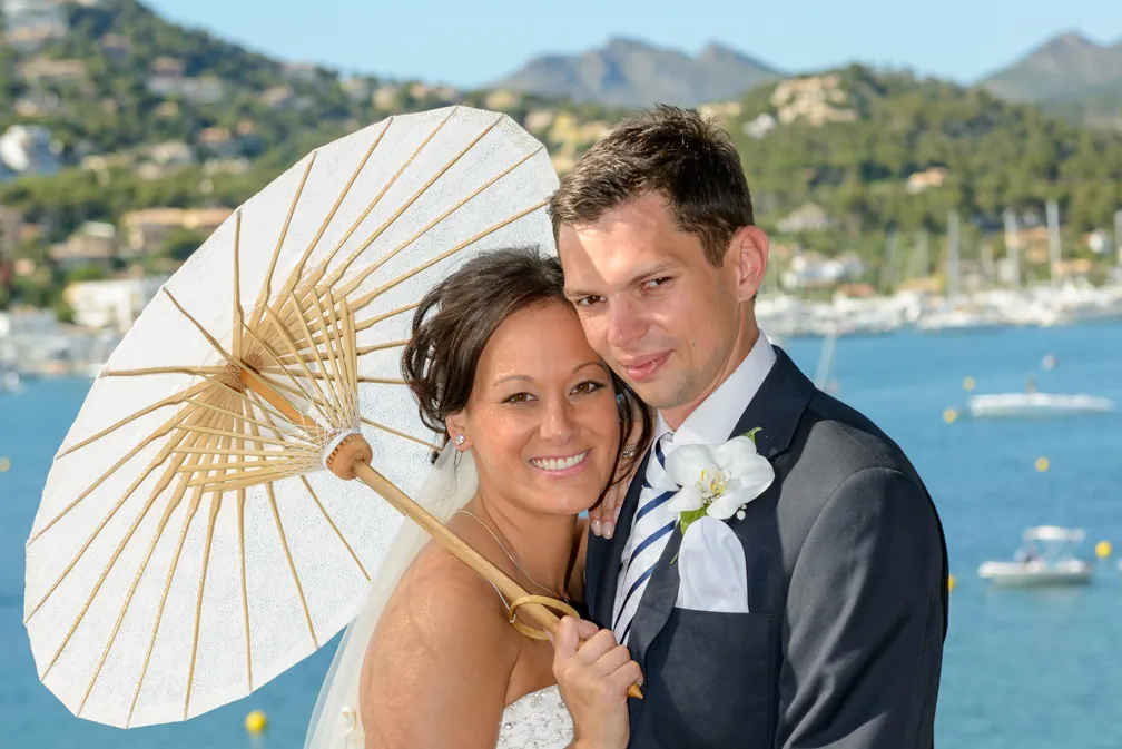 Smiling bride and groom embracing outdoors near water with boats and hills in the background, bride holding a white parasol.