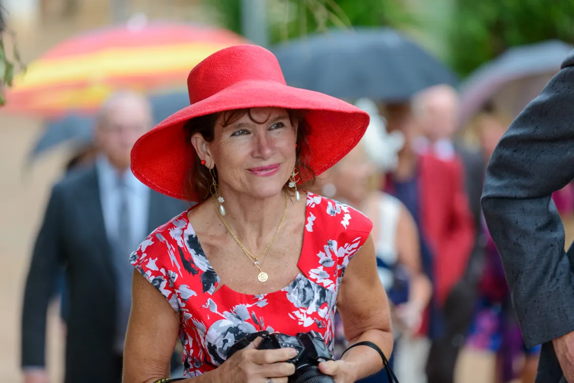 Woman in a red wide-brimmed hat and floral dress holding a camera, smiling with blurred people in the background.