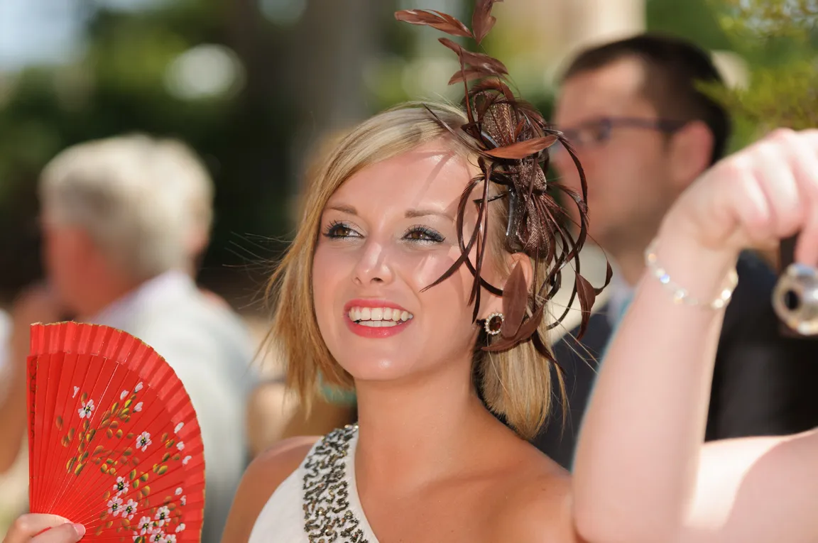 Smiling woman in a white dress with decorative beads wearing a brown feathered fascinator and holding a red floral hand fan.