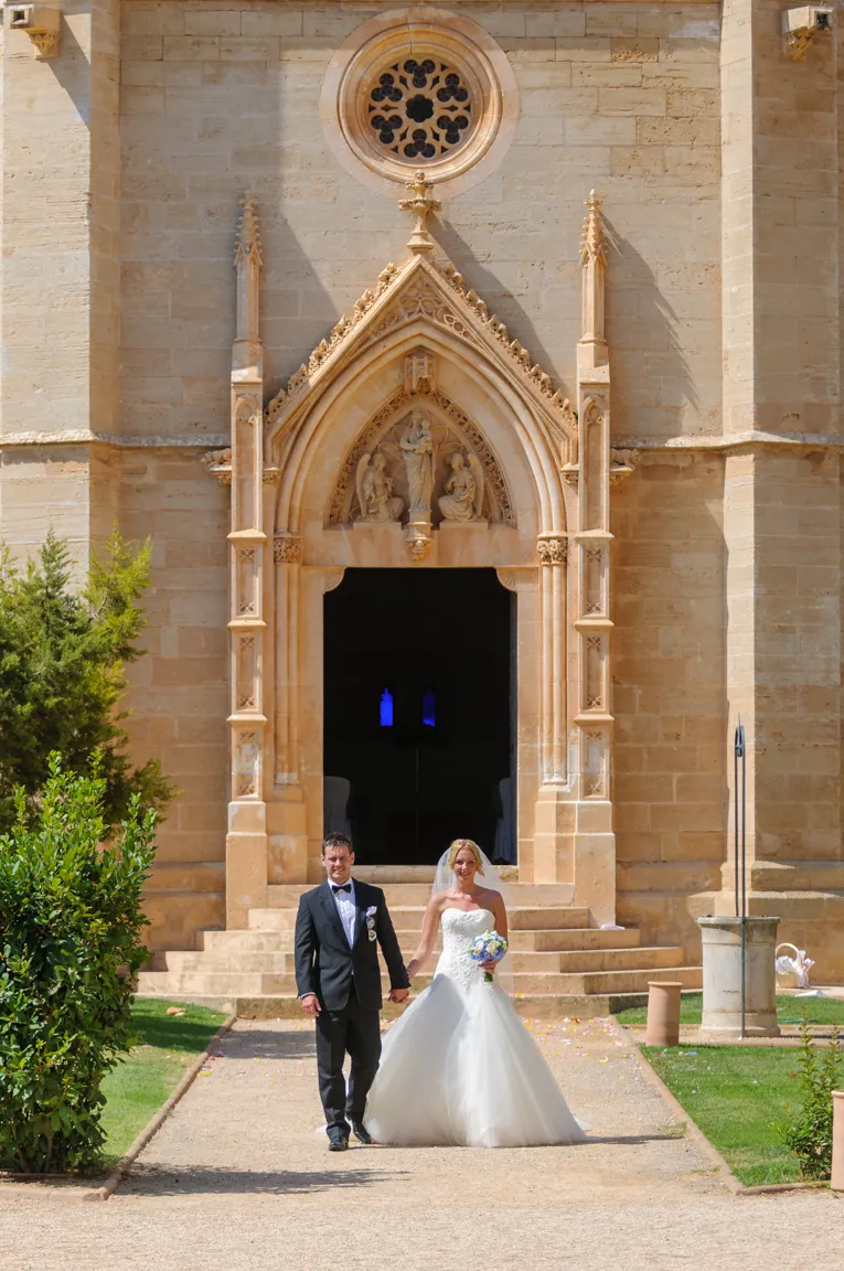 Bride in white wedding gown and groom in black tuxedo holding hands and walking outside a stone church entrance with gothic arch and sculptures.