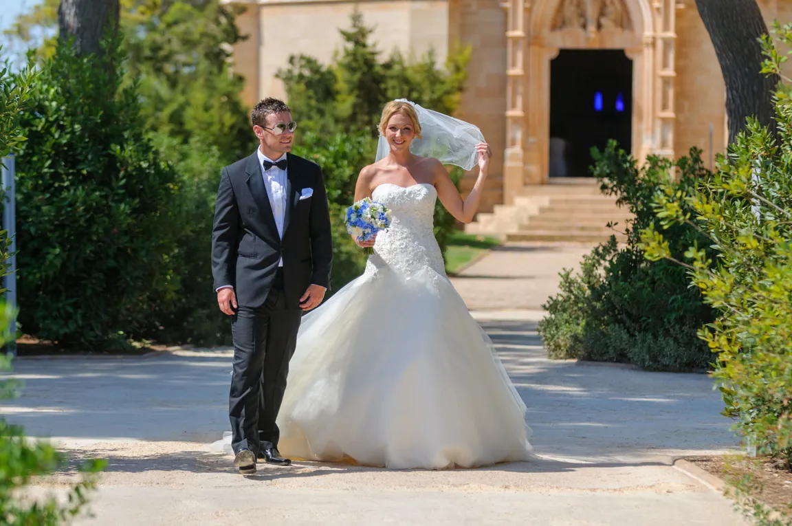 Bride in a strapless white wedding gown holding a bouquet and adjusting her veil, walking beside a groom in a black tuxedo with sunglasses, in an outdoor garden path with a stone building in the background.