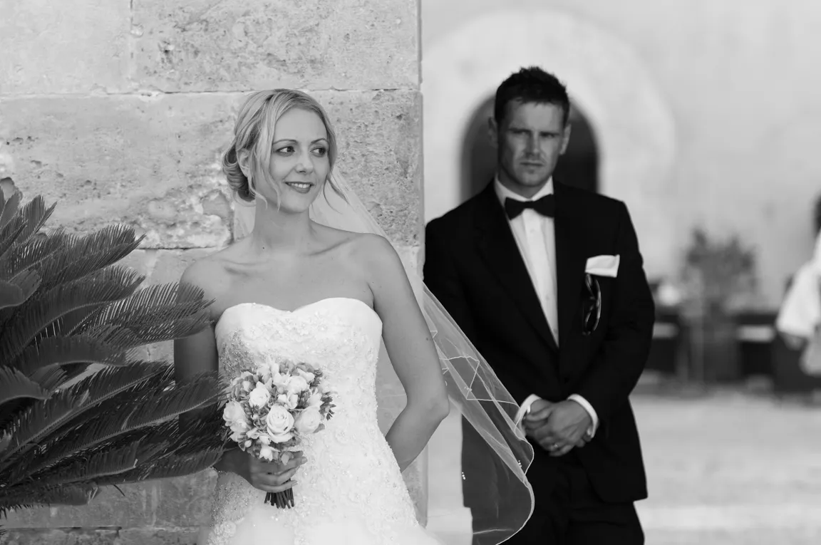 Bride in a strapless wedding dress holding a bouquet, standing by a stone wall, with a groom in a tuxedo blurred in the background.