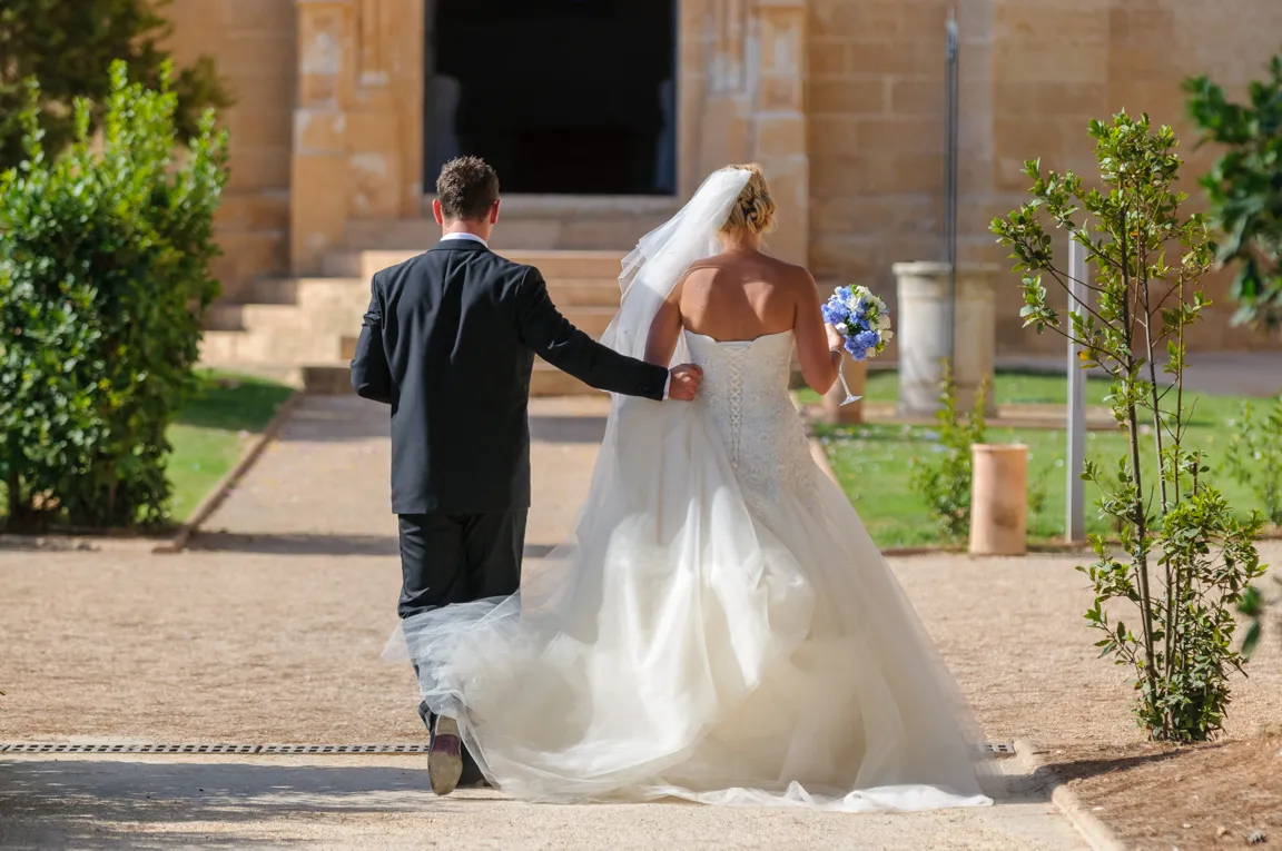 Bride in white gown holding a blue and white bouquet walking arm in arm with a man in a black suit toward a stone building.