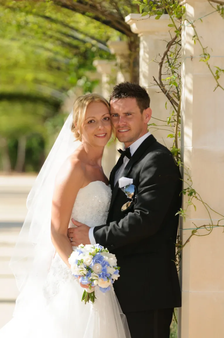Bride and groom embracing outdoors under a green leafy arch, the bride holding a bouquet of white and blue flowers.