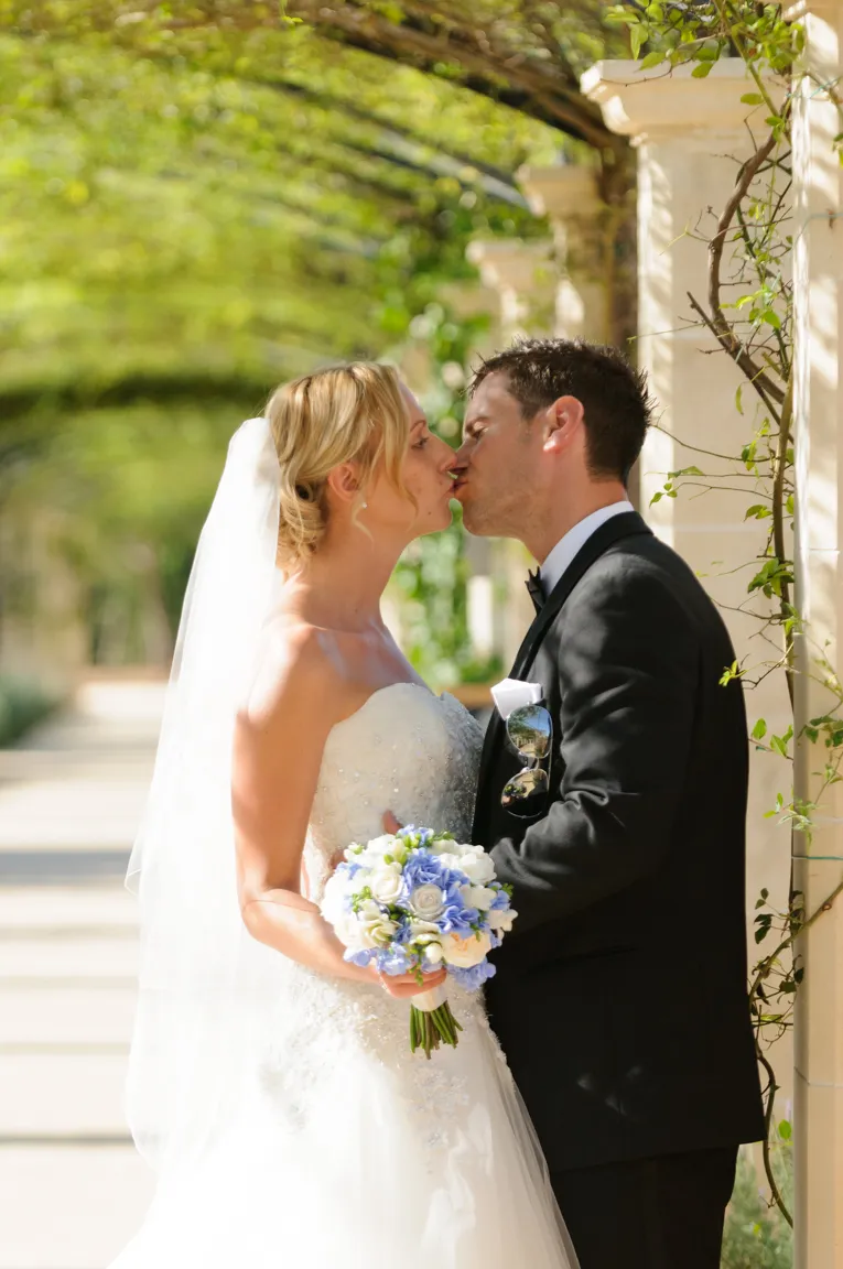Bride and groom sharing a kiss outdoors with bride holding a bouquet of white and blue flowers.