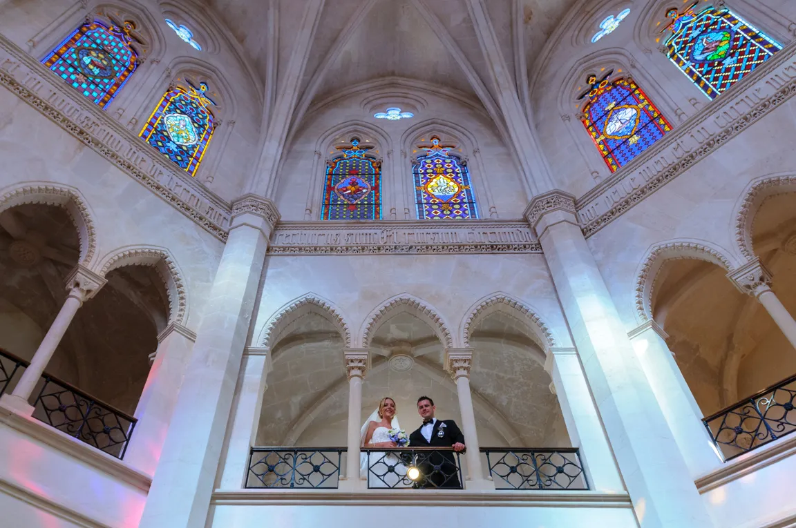 Bride and groom standing on ornate balcony inside a grand stone building with stained glass windows above.