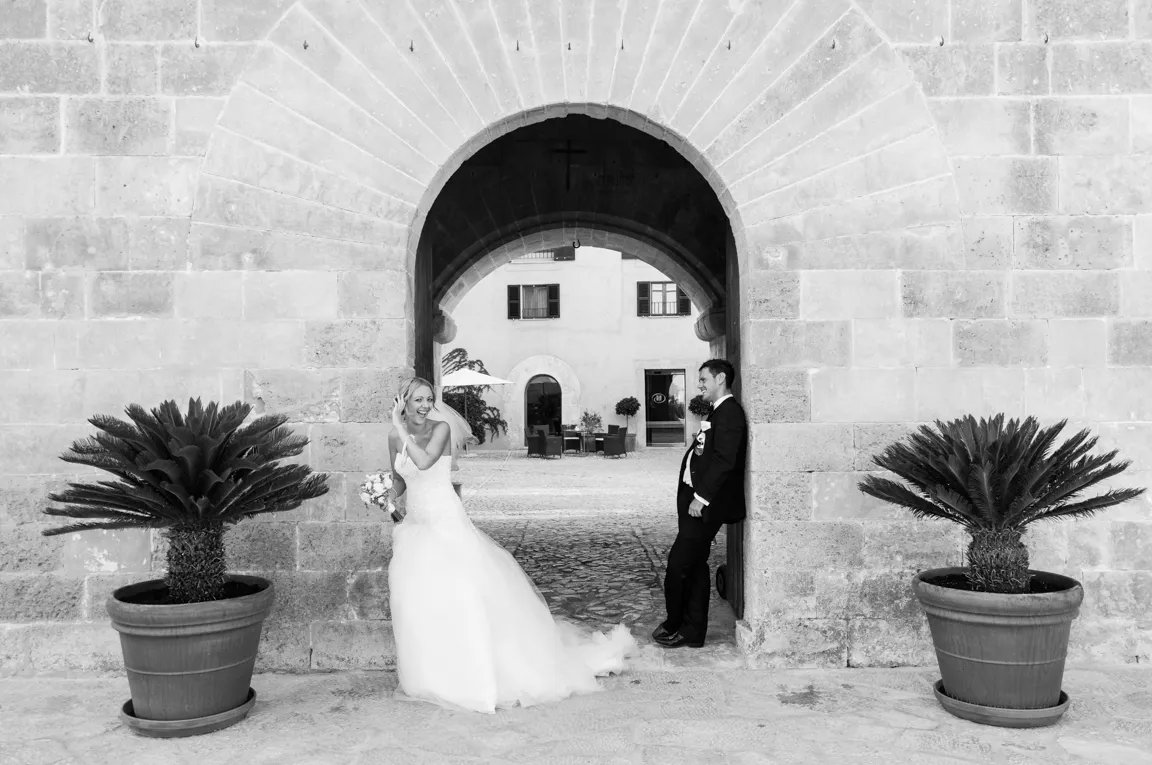 Bride and groom smiling and posing under a stone archway with potted plants on each side in black and white.
