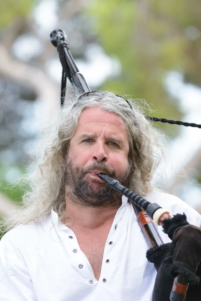 Man with long curly gray hair playing the bagpipes outdoors against a blurred natural background.