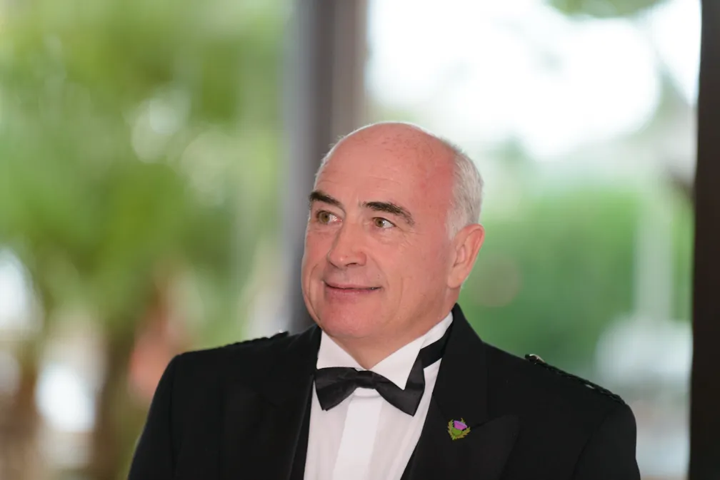 Smiling bald man in a black tuxedo with a bow tie and a flower pin on the lapel, standing outdoors with blurred greenery in the background.