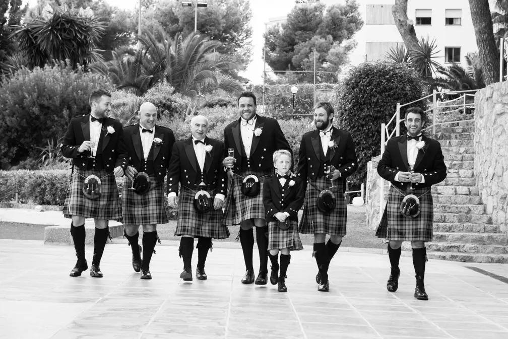 Seven men and a boy wearing traditional kilts and tuxedo jackets walking outdoors, some holding drinks and smiling.