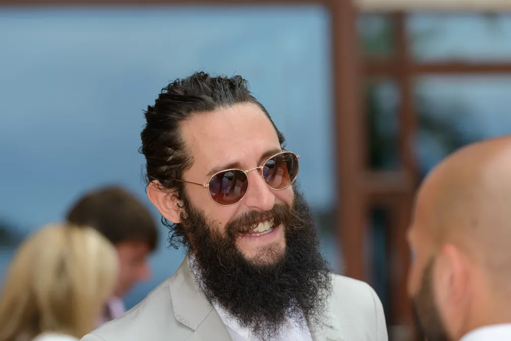 Man with long dark beard, tied-back hair, and sunglasses smiling while talking to another person outdoors.