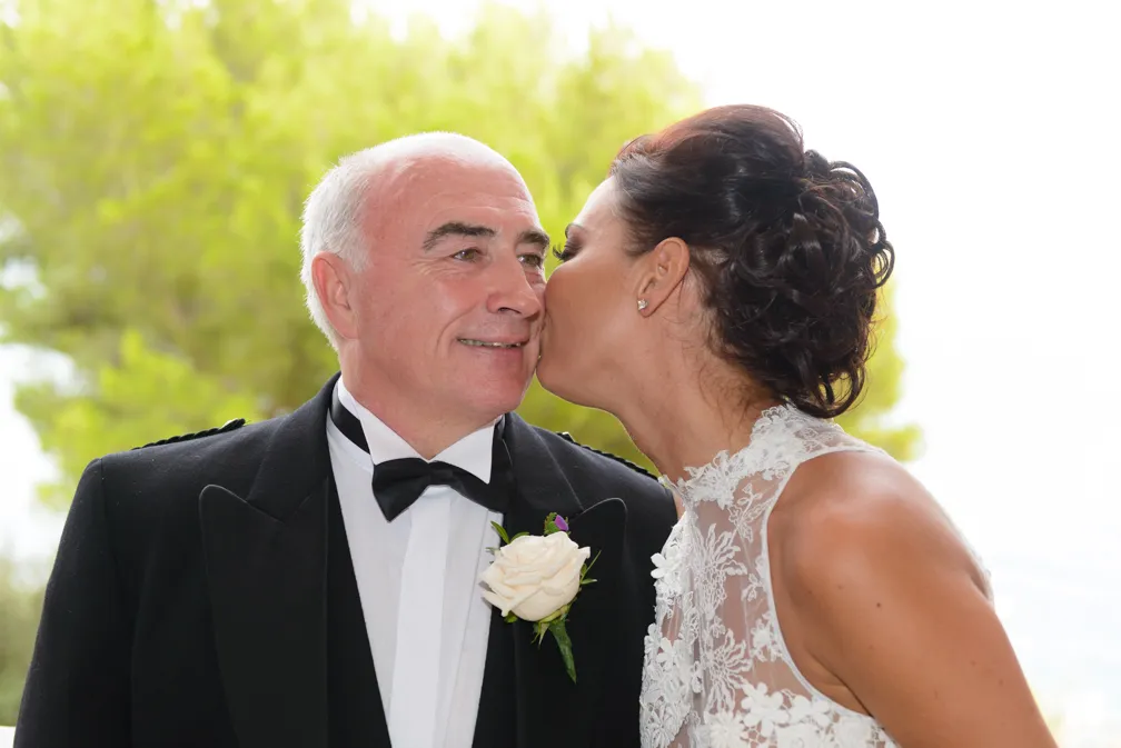 Bride in lace dress kissing an older man in a tuxedo with a white rose boutonniere on the cheek outdoors.