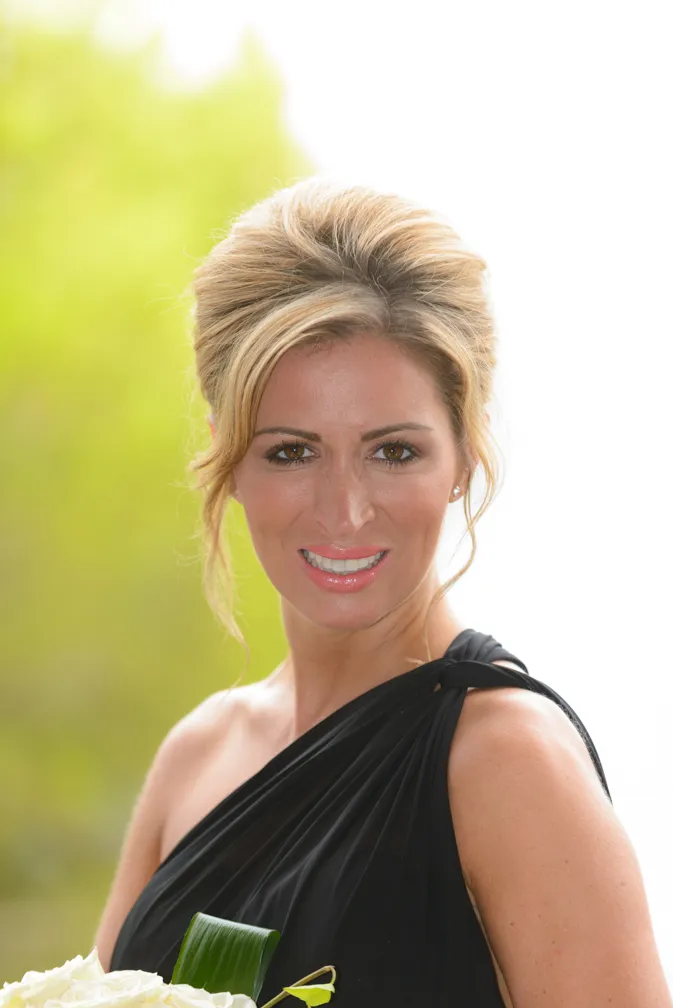 Smiling woman with blonde hair styled up wearing a black one-shoulder dress holding white flowers with green foliage.