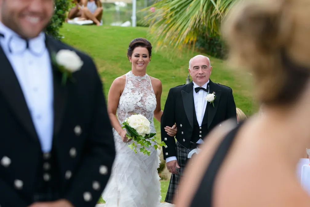Bride in a white lace gown holding a bouquet of white roses walks arm-in-arm with an older man dressed in a traditional black tuxedo with a kilt outdoors on a grassy slope.