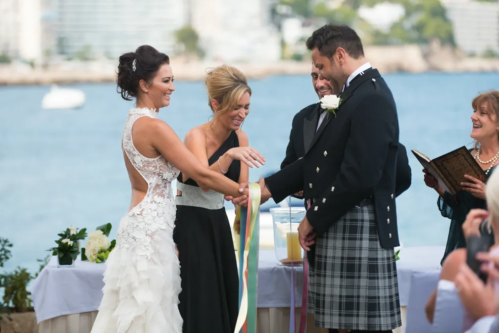 Bride, groom in a kilt, and bridesmaid smiling and holding colorful ribbons during a beachside wedding ceremony.