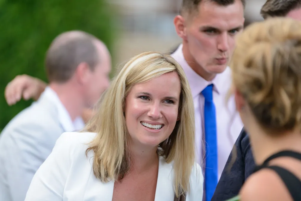 Smiling blonde woman in a white blazer talking with others in a social outdoor setting.