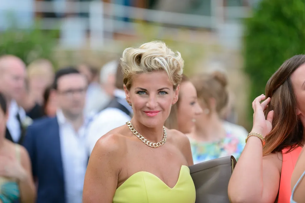 Smiling blonde woman in a strapless yellow dress wearing a gold chain necklace at an outdoor event.