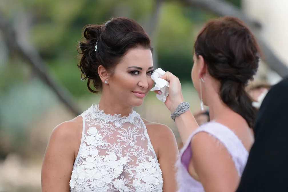 Woman in white lace dress smiling as another woman wipes her tears with a tissue.