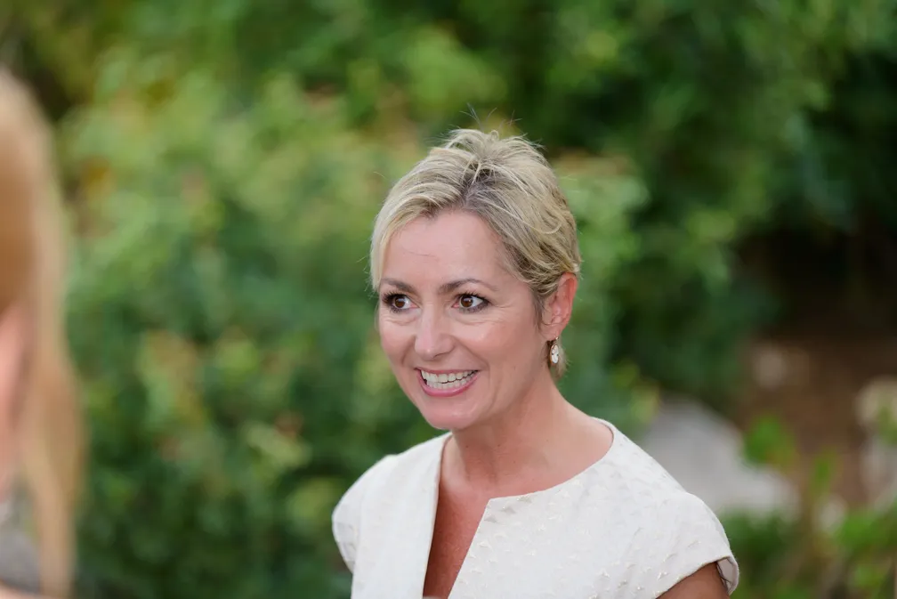 Smiling woman with short blonde hair and white sleeveless top outdoors with greenery background.