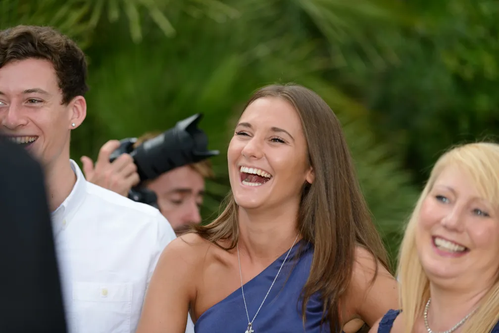 Three people laughing and smiling outdoors, with green foliage in the background.