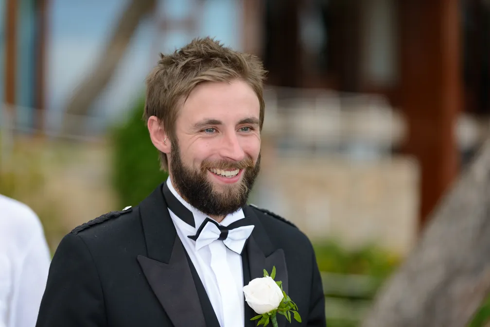 Smiling man with beard wearing a black tuxedo with white bow tie and white rose boutonniere.