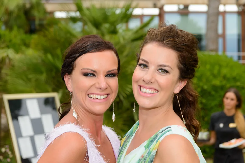 Two smiling women dressed in formal attire posing outdoors with greenery and a blurred waiter in the background.
