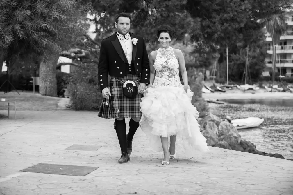 A man in a traditional Scottish kilt and a woman in a white wedding dress walking together outdoors near water.