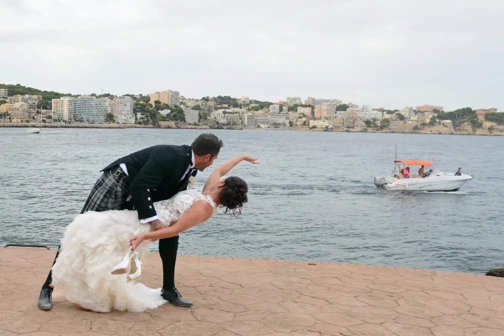 Bride in a white wedding dress holding her shoes as the groom in a black jacket and kilt dips her near a waterfront with a boat and cityscape in the background.