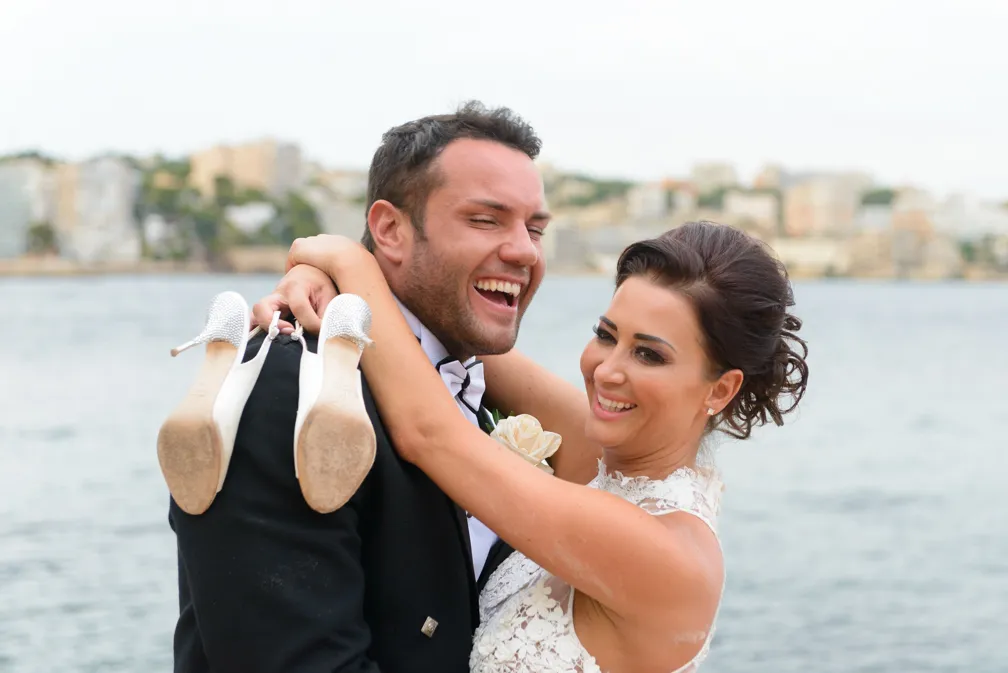 Smiling bride holding her white high-heeled shoes over the shoulder of a laughing groom in a black tuxedo by the water.