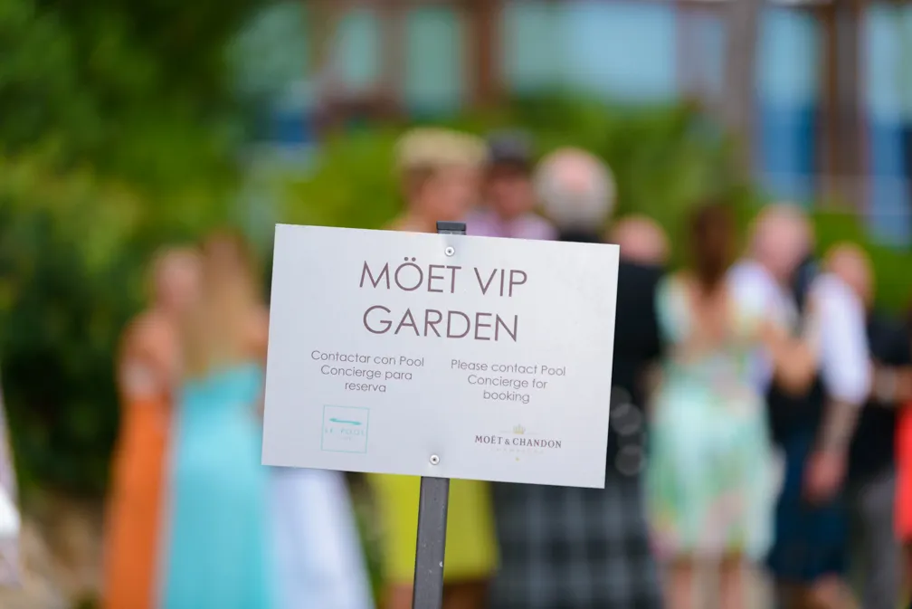 Sign reading 'MOËT VIP GARDEN' with instructions to contact pool concierge for booking, with blurred people in colorful clothing in the background.