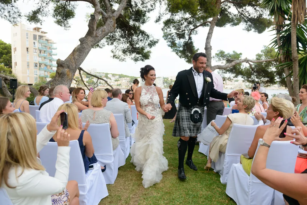 Bride and groom holding hands and smiling as they walk down the outdoor aisle surrounded by seated guests during their wedding ceremony.
