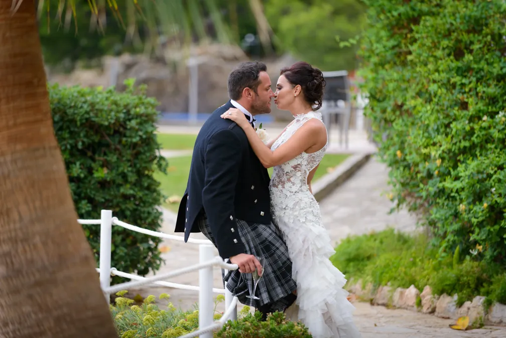 Bride in a lace wedding dress and groom in a traditional Scottish kilt sharing a kiss outdoors by greenery.