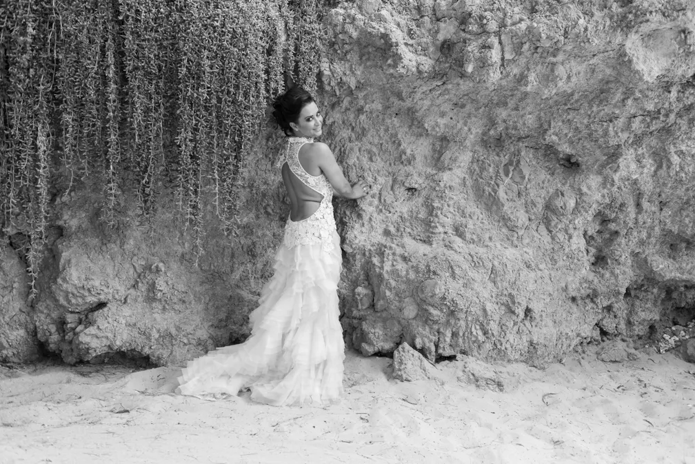 Woman in a white backless wedding dress standing on sand, leaning against a textured rock wall with hanging vegetation.