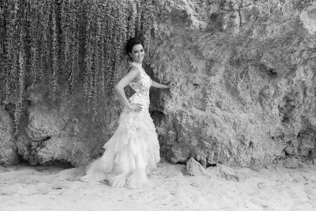 Bride in a layered wedding dress posing with one hand on her hip and the other on a rocky cliff with hanging foliage.