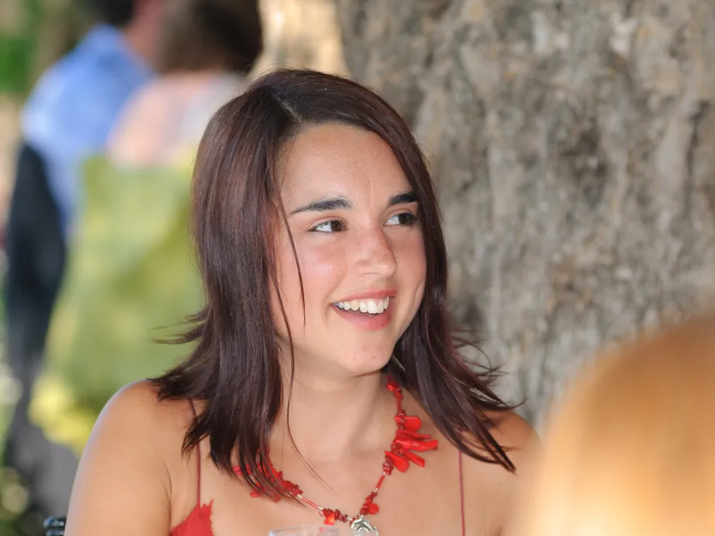 Smiling woman with dark hair wearing a red necklace and a red top, sitting outdoors near a tree.