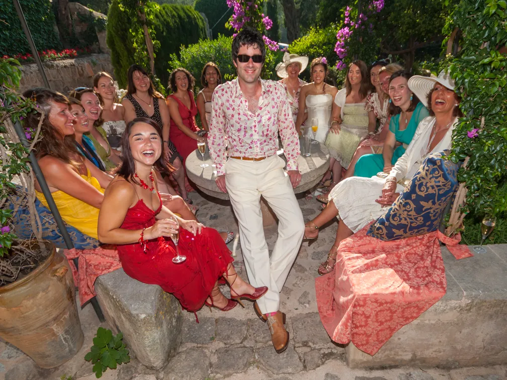Group of people dressed in colorful summer clothing smiling and sitting around an outdoor stone table in a garden setting.