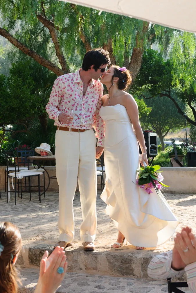 Bride in white dress kissing groom in floral shirt and white pants while holding hands and bouquet outdoors.