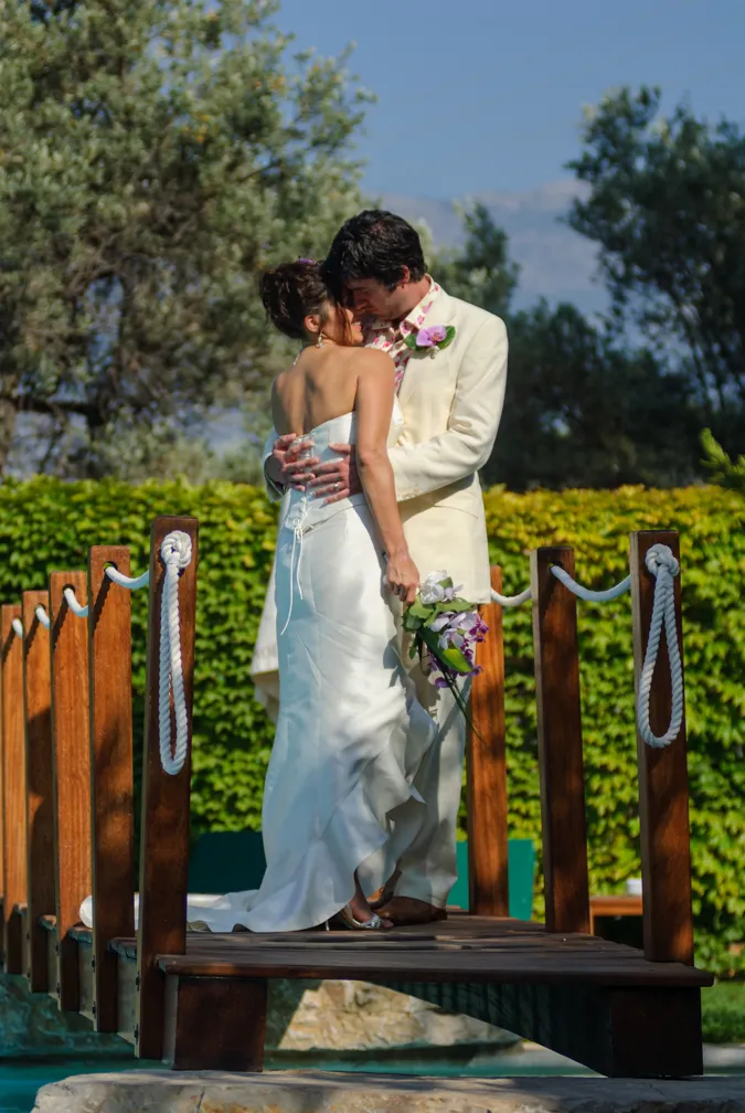 Bride and groom embracing on a wooden bridge outdoors with greenery and mountains in the background.