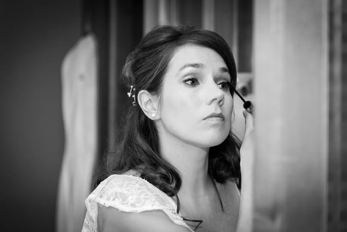 Woman with dark hair and lace dress applying mascara in front of a mirror.