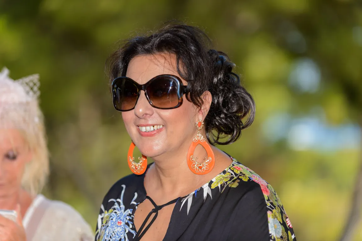 Smiling woman with dark curly hair wearing large sunglasses and orange decorative earrings in an outdoor setting.