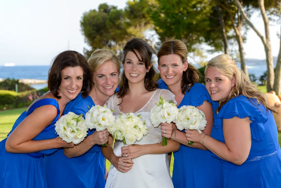 Bride in white dress with four bridesmaids in blue dresses holding white flower bouquets, posing outdoors near the sea.