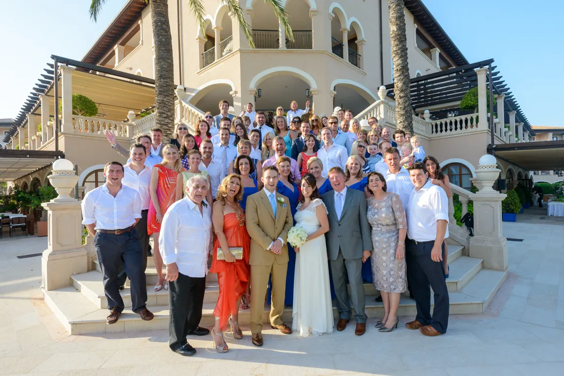 Large group of people posing on steps in front of a Mediterranean-style building, with a bride in a white dress holding a bouquet and a groom in a tan suit at the center.