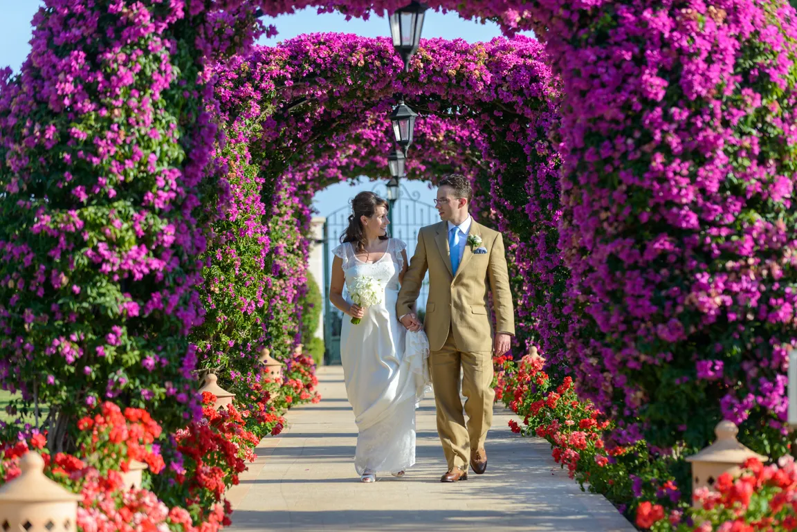 Bride and groom walking hand in hand under vibrant pink floral arches on a sunny day.